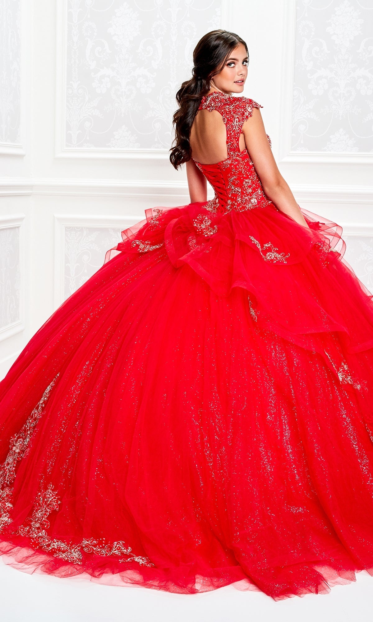 A woman wears the Princesa Beaded Lace Quinceañera Dress PR11923, a vibrant red gown with beaded lace and a full tulle skirt, facing slightly backward in an ornate white room.